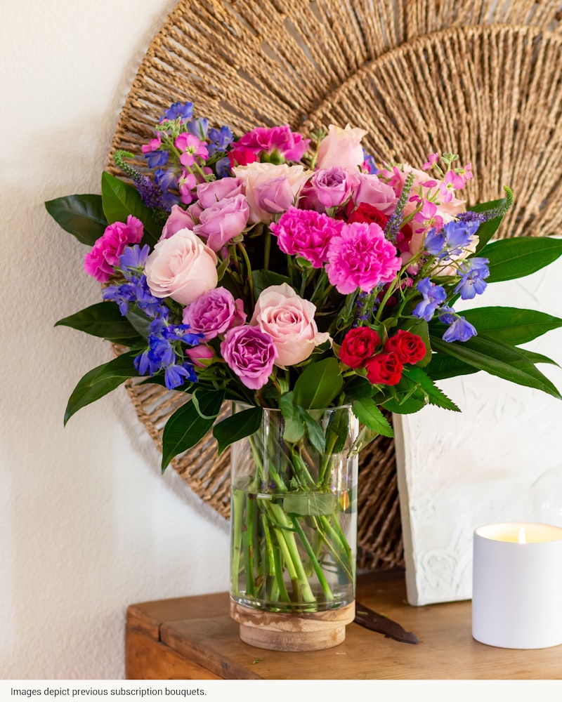 Lush bouquet of pink roses, purple flowers, and green foliage in a clear glass vase on a wooden table with a lit candle and woven wall decor in the background.