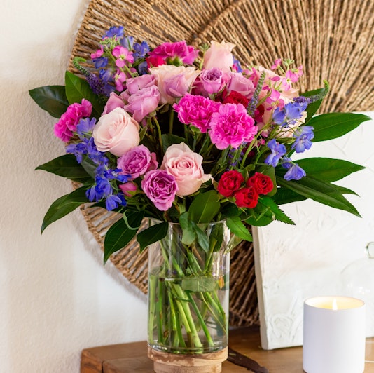 Vibrant mixed bouquet featuring pink roses, lavender blooms, and lush greens in a glass vase.