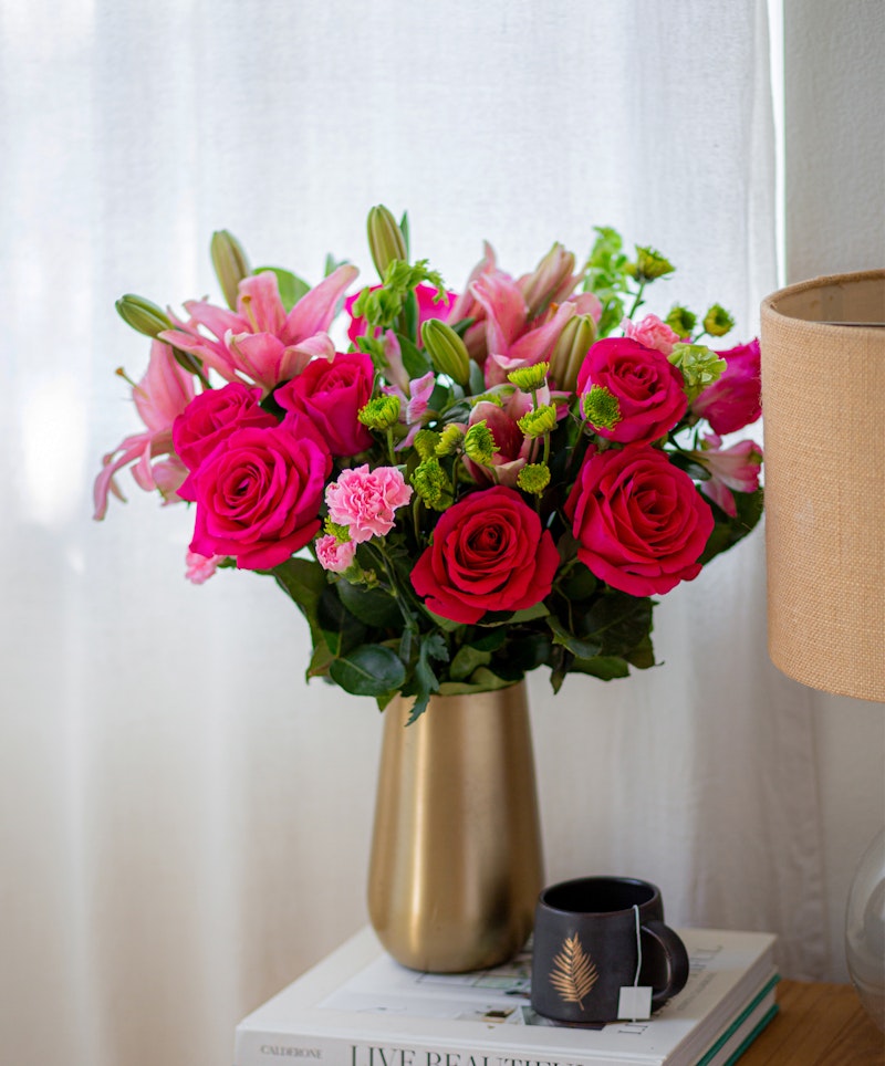 Vibrant bouquet of pink lilies and red roses in a gold vase on a stack of books next to a coffee mug, with a textured lamp in a cozy interior setting.