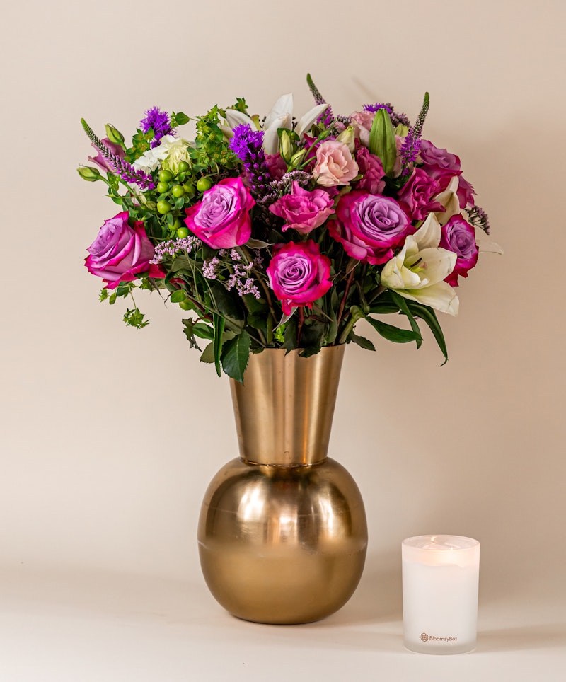 Vibrant bouquet of purple and pink flowers, including roses and lilies, placed in a metallic gold vase beside a white lit candle on a neutral background.