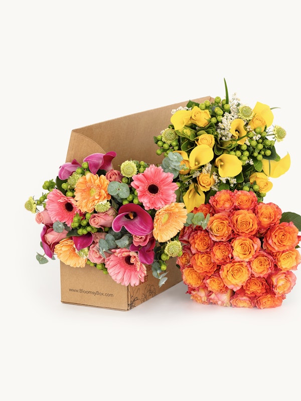 A vibrant assortment of fresh flowers including pink gerberas, orange roses, and purple calla lilies in a cardboard flower box on a white background.