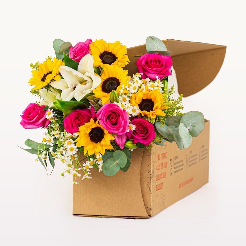 Vibrant flower arrangement with sunflowers, pink roses, and lilies protruding from an opened cardboard box on a white background, symbolizing a fresh floral delivery.