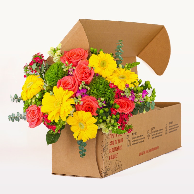 A vibrant bouquet of yellow and pink flowers, including roses and gerberas, artistically arranged in an eco-friendly brown cardboard box on a white background.