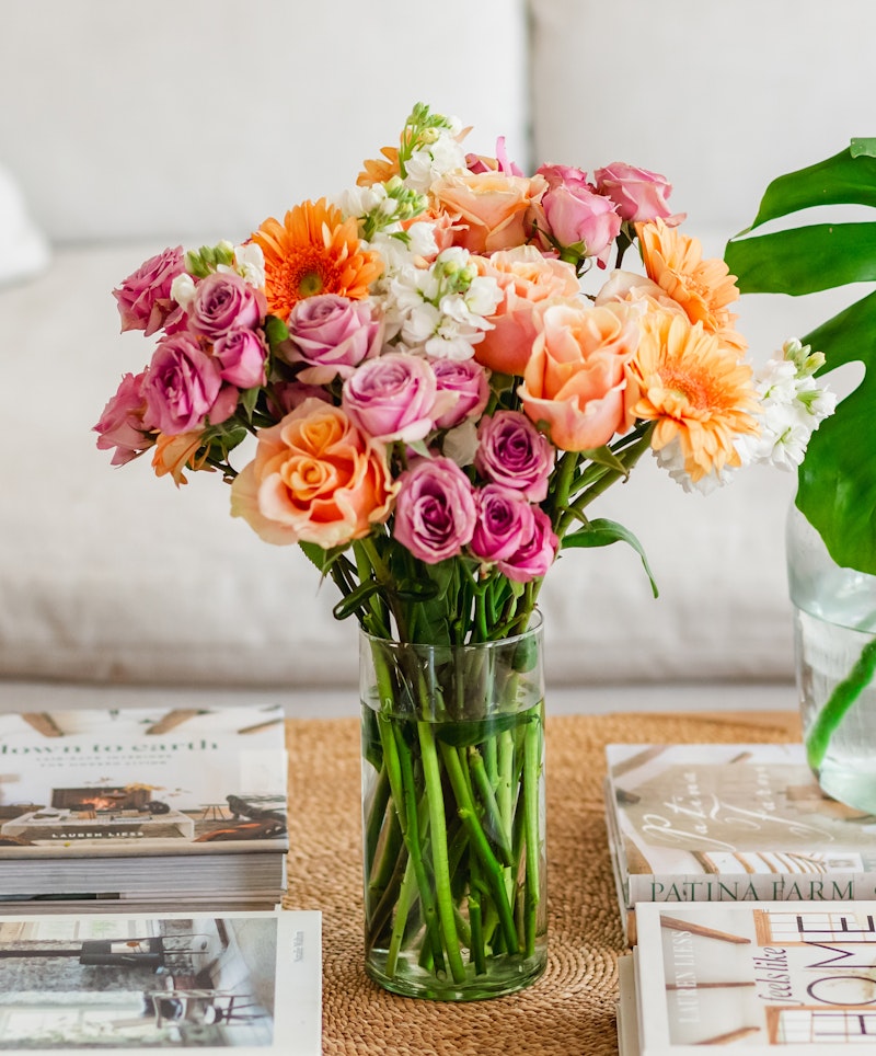 Vibrant bouquet of fresh flowers with roses, lilies, and daisies in shades of orange, pink, and white arranged in a clear glass vase on a table with magazines.