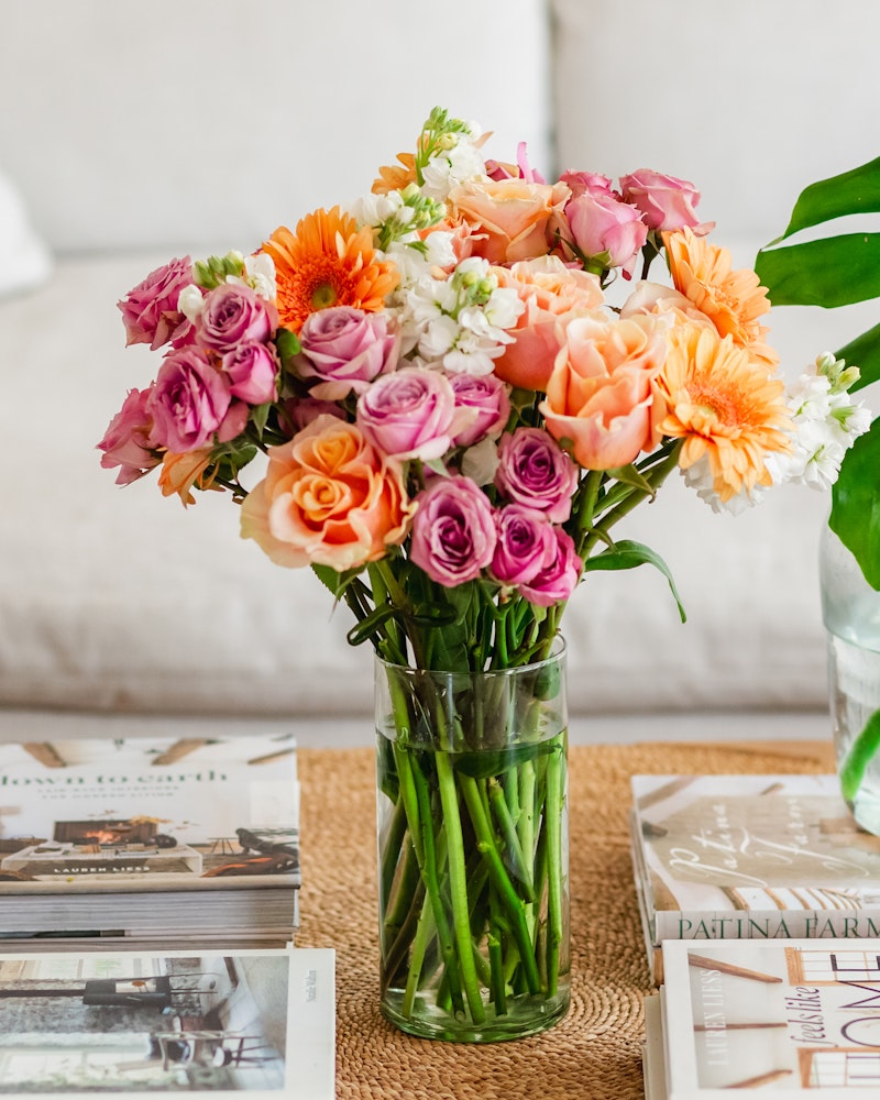 Vibrant bouquet of fresh flowers with roses, lilies, and daisies in shades of orange, pink, and white arranged in a clear glass vase on a table with magazines.