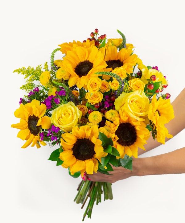 A vibrant bouquet of yellow sunflowers and roses with touches of purple flowers and greenery held by a person against a white background, showcasing a fresh floral arrangement.