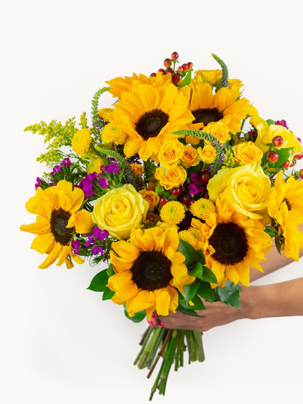 A vibrant bouquet of yellow sunflowers and roses with touches of purple flowers and greenery held by a person against a white background, showcasing a fresh floral arrangement.