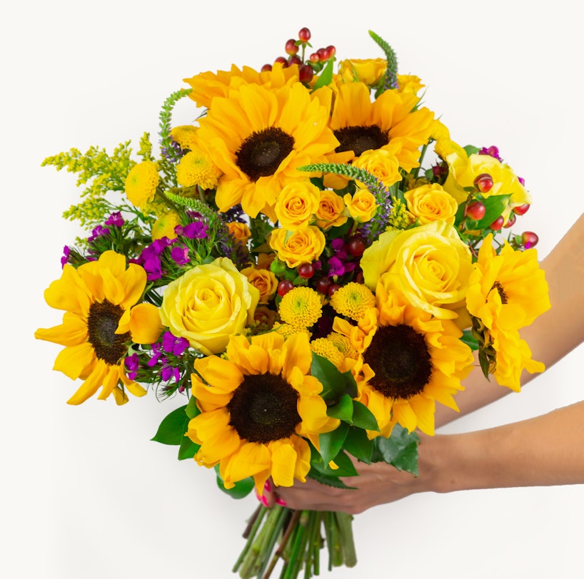 A vibrant bouquet of yellow sunflowers and roses with touches of purple flowers and greenery held by a person against a white background, showcasing a fresh floral arrangement.