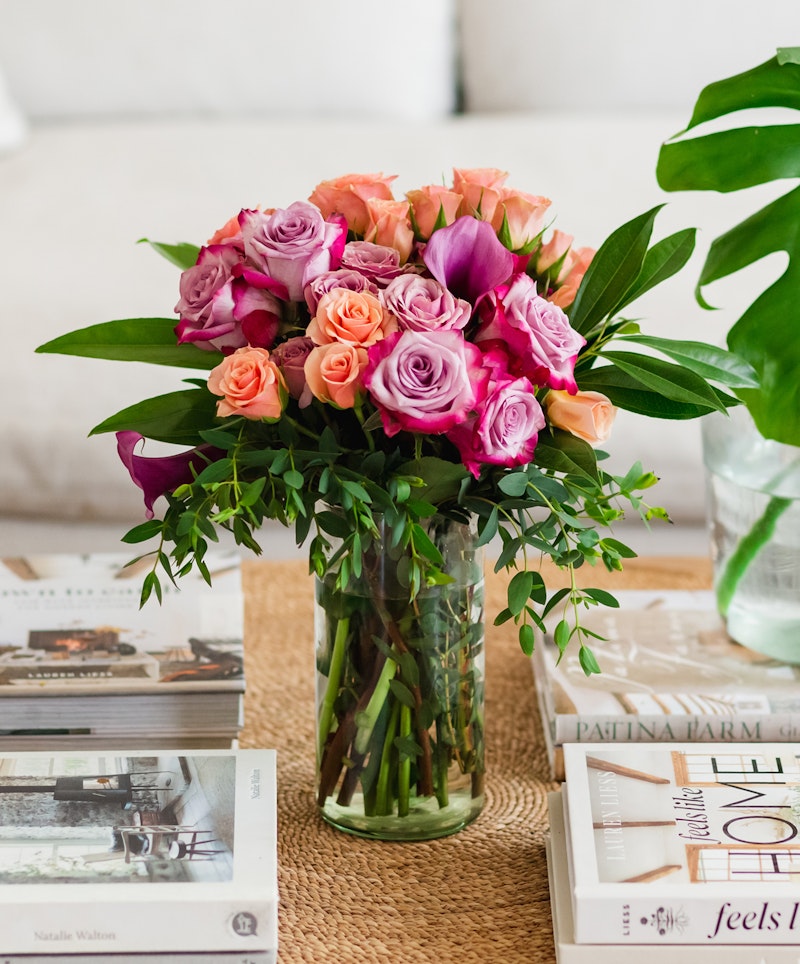 Vibrant bouquet of pink and orange roses with lush greenery in a clear glass vase on a table adorned with stylish coffee table books, exuding a cozy home atmosphere.