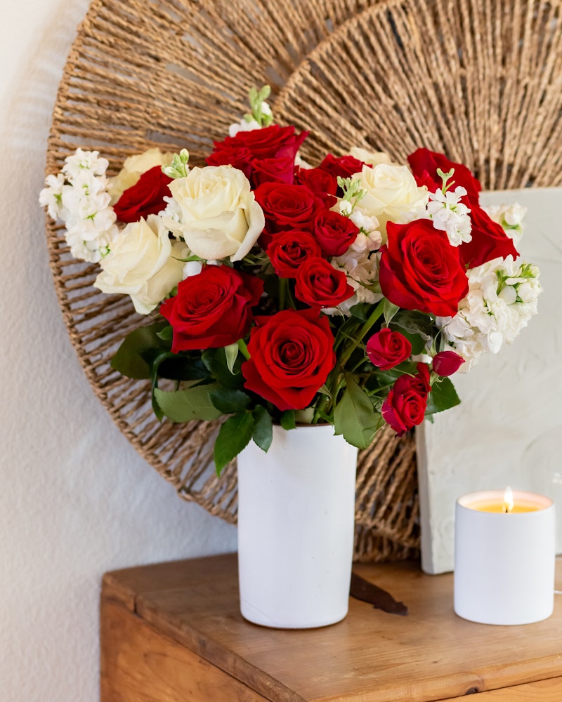 Bouquet of vibrant red roses and delicate white flowers in a white vase on a wooden table, with a lit candle and a woven circular backdrop.