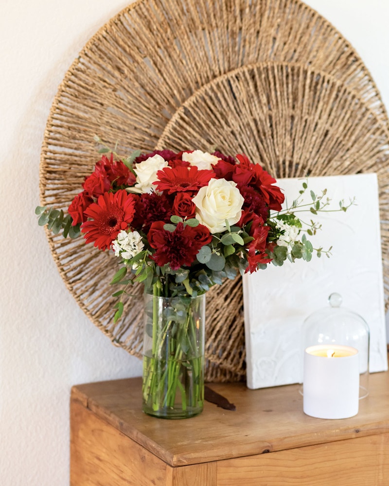 A vibrant bouquet of red and white flowers in a glass vase on a wooden table, with a woven circular wall decor in the background and a lit candle in the foreground.