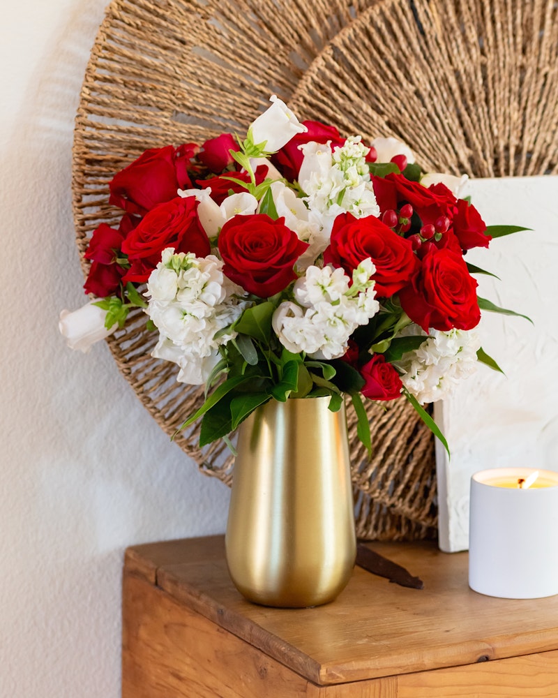 Vibrant bouquet of red roses and white flowers in a gold vase on a wooden table, with a lit candle and decorative woven wall art in the background.