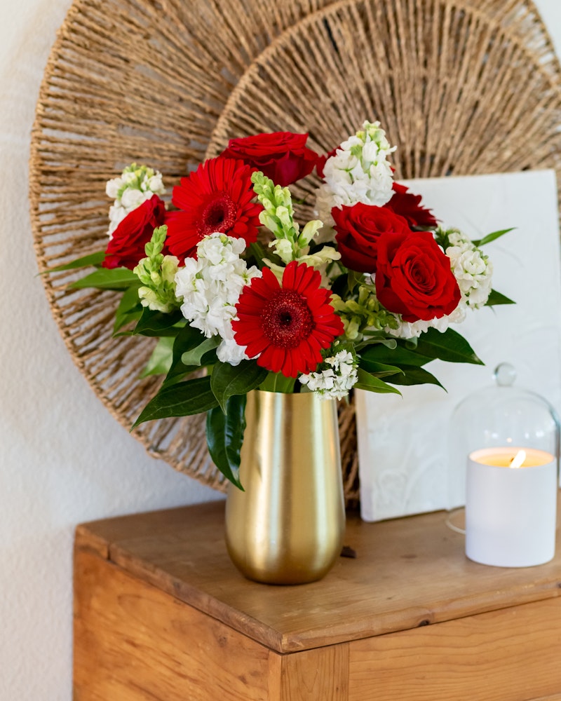 Vibrant red gerberas and roses arranged with white blooms in a golden vase on a wooden table, with a lit candle and wicker decor in the background.