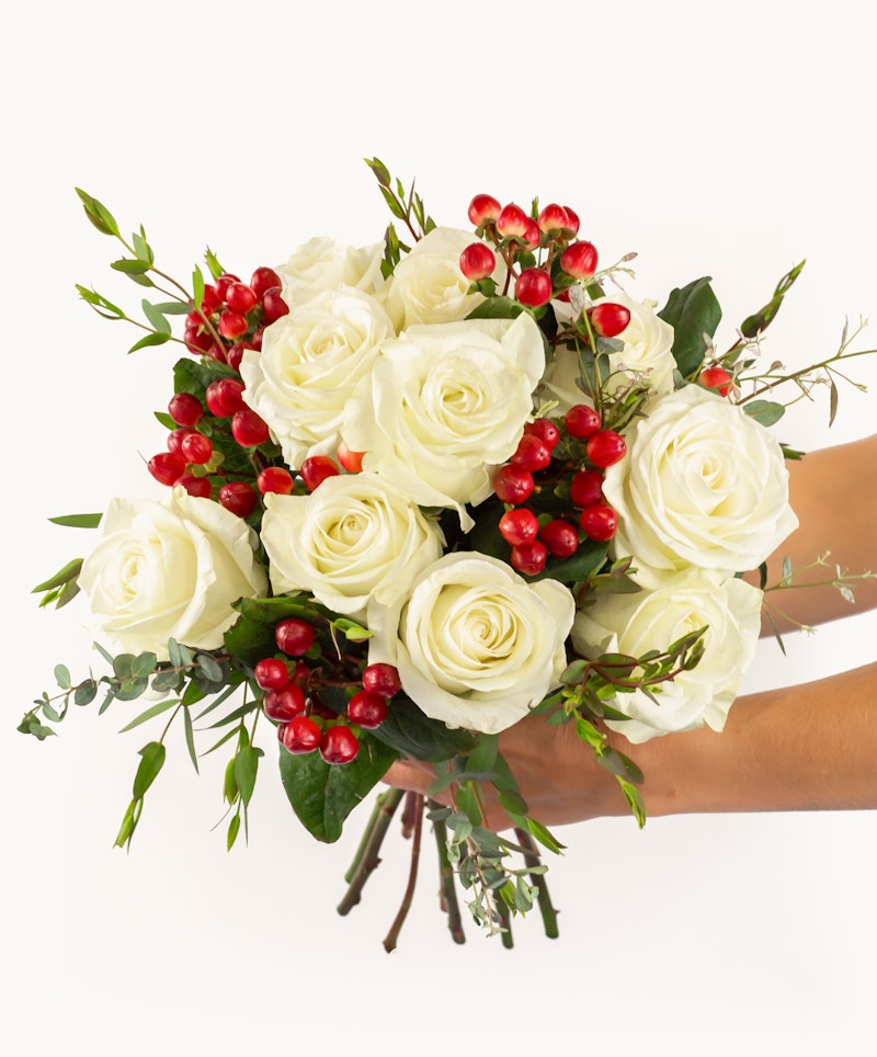 A person holding a bouquet of white roses accented with red berries and green foliage against a clean white background, perfect for special occasions.