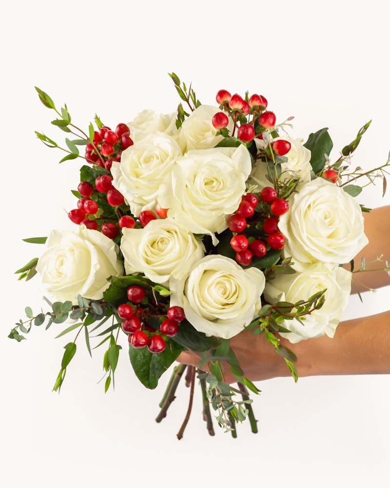 A person holding a bouquet of white roses accented with red berries and green foliage against a clean white background, perfect for special occasions.