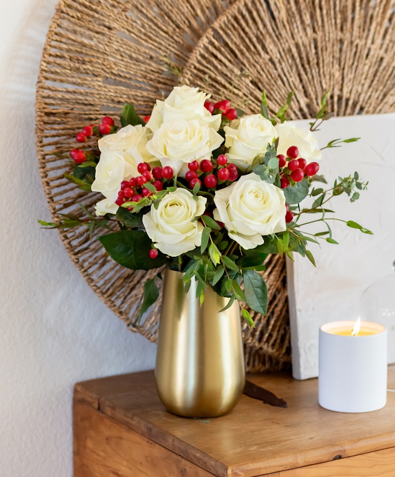A vibrant bouquet of white roses and red berries in a golden vase, set against a rustic backdrop with a woven round decoration and a lit candle on a wooden surface.