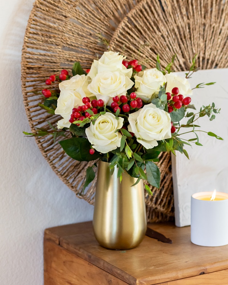 A vibrant bouquet of white roses and red berries in a golden vase, set against a rustic backdrop with a woven round decoration and a lit candle on a wooden surface.