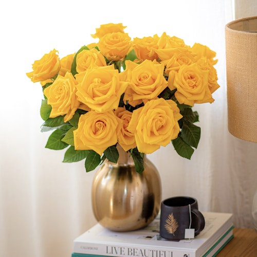 Vibrant bouquet of yellow roses in a metallic gold vase, placed on books beside a mug and lamp, with a backdrop of a white wall.