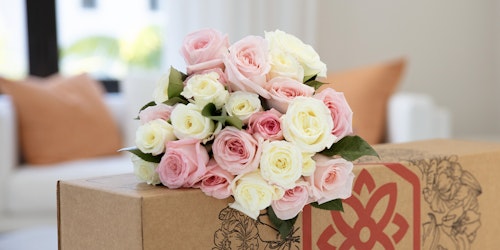 Beautiful bouquet of pink and white roses atop a decorated cardboard box, set against a modern living room with white walls and orange pillows.