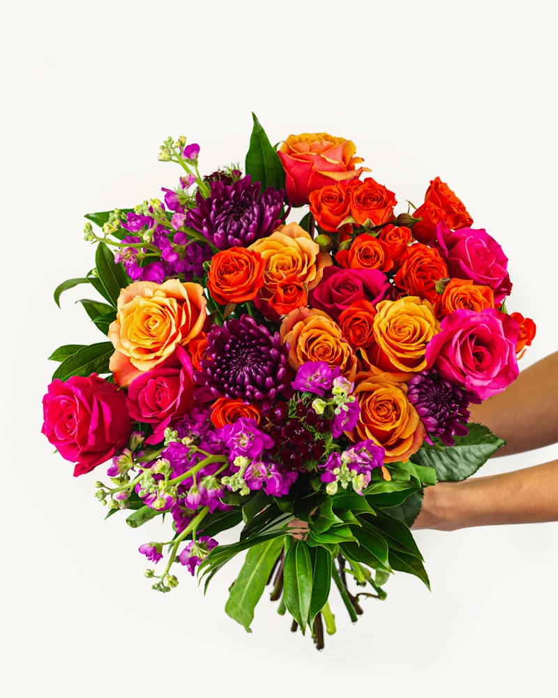 Vibrant bouquet of flowers featuring a mix of orange, pink, and purple blooms with lush green leaves, held against a white background.