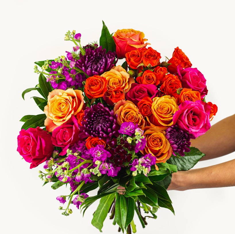 Vibrant bouquet of flowers featuring a mix of orange, pink, and purple blooms with lush green leaves, held against a white background.