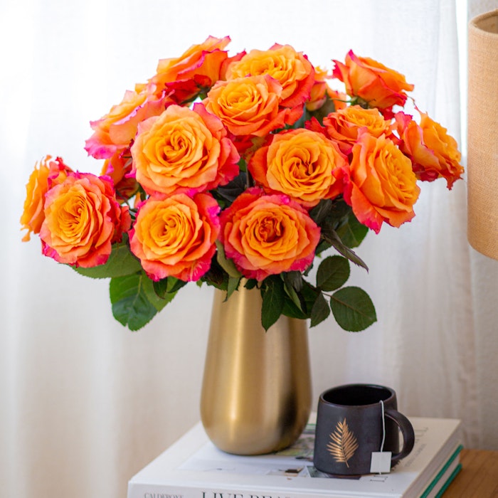 A vibrant bouquet of orange roses in a gold vase on a table with books and a black coffee mug, beside a softly lit lamp, adding warmth and color to the room.