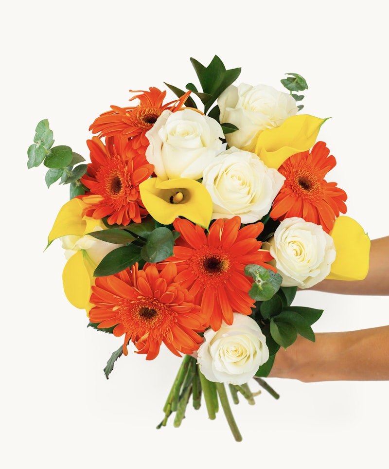 Vibrant bouquet of flowers featuring orange gerberas, white roses, and yellow lilies with green leaves, held by a hand against a white background.