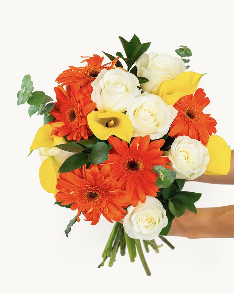 Vibrant bouquet of flowers featuring orange gerberas, white roses, and yellow lilies with green leaves, held by a hand against a white background.