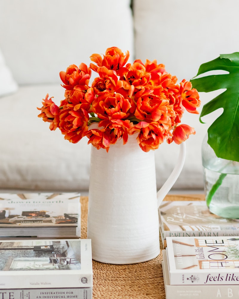 Vibrant orange tulips arranged in a white vase on a coffee table with assorted magazines and a green monstera leaf, adding a splash of color to a cozy living room setting.