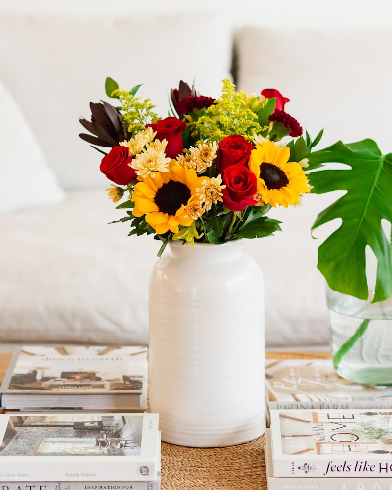 Bright bouquet of sunflowers, red roses, and various blooms in a white vase on a table with books, conveying a cozy, welcoming home atmosphere.