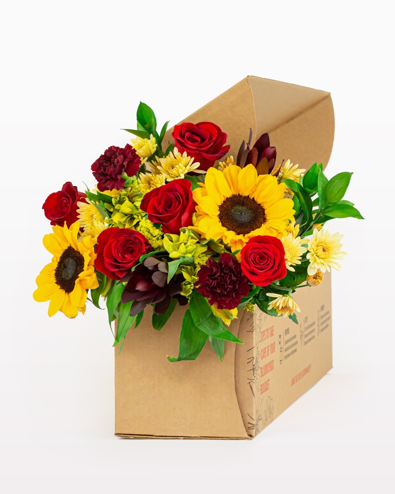 Bouquet of fresh flowers including sunflowers, red roses, and chrysanthemums arranged in a brown cardboard flower delivery box against a white background.