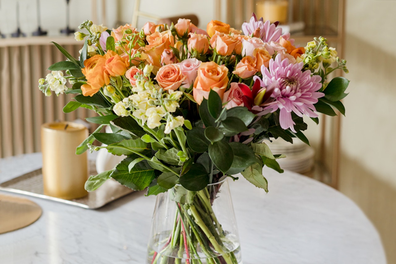 Vibrant floral arrangement featuring orange roses, pink daisies, and lush greenery in a vase.