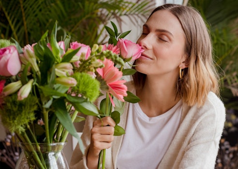 Woman in a cozy white sweater enjoying the scent of a pink flower with a bouquet of fresh flowers in the background, evoking a sense of spring and relaxation.