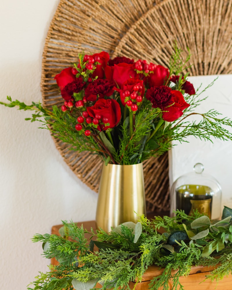 Vibrant red flowers and green foliage arranged in a gold vase, with additional greenery spread on the wooden surface, set against a white wall with a circular wicker decoration.