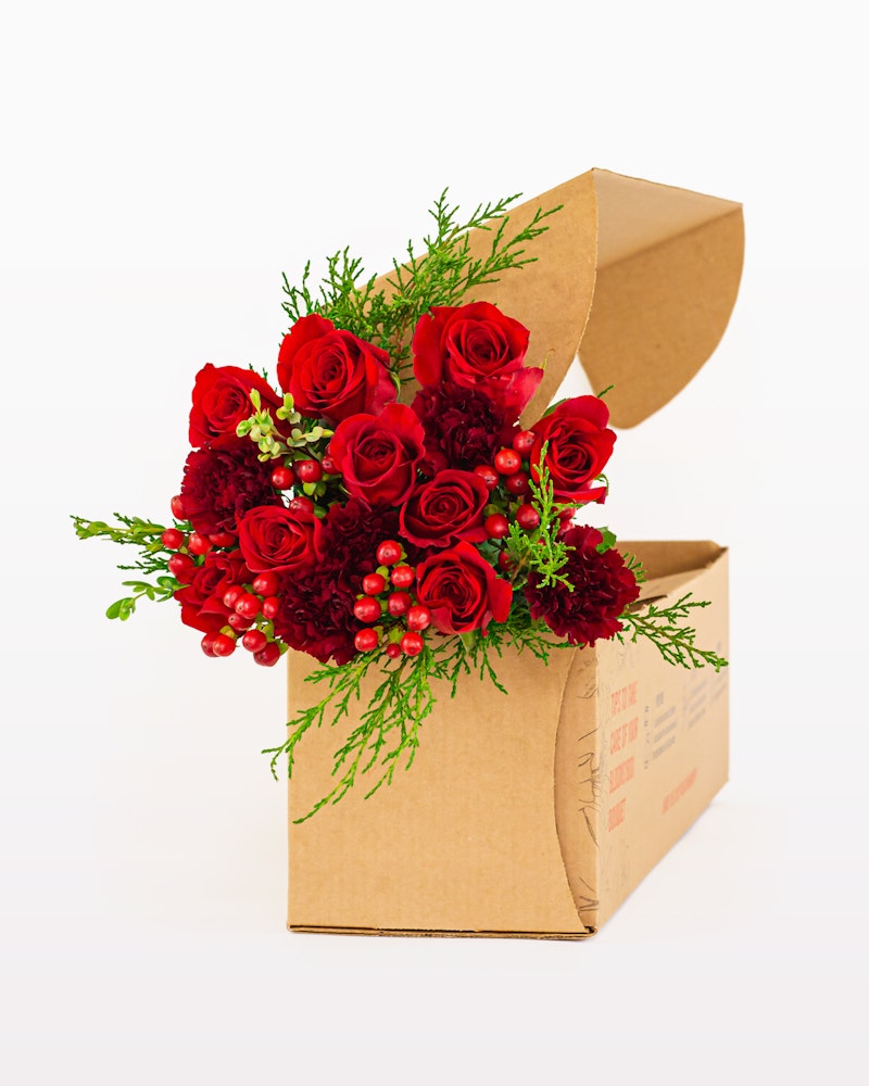 Bouquet of red roses and burgundy flowers with green fern-like foliage arranged in an open cardboard gift box on a white background, symbolizing a special delivery.