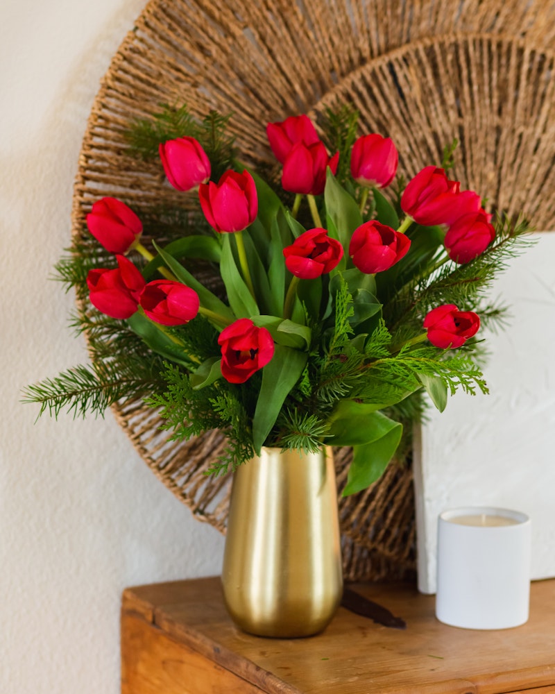 A vibrant bouquet of red tulips arranged in a golden vase on a wooden table, complemented by greenery and a woven wall decoration, with a white candle nearby.