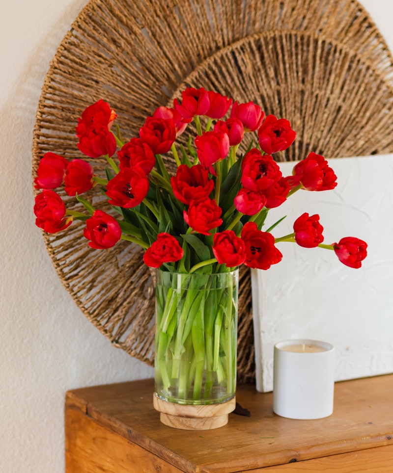Vibrant red tulips arranged in a clear glass vase on a wooden side table, with decorative woven wall art in the background and a white candle to the side.