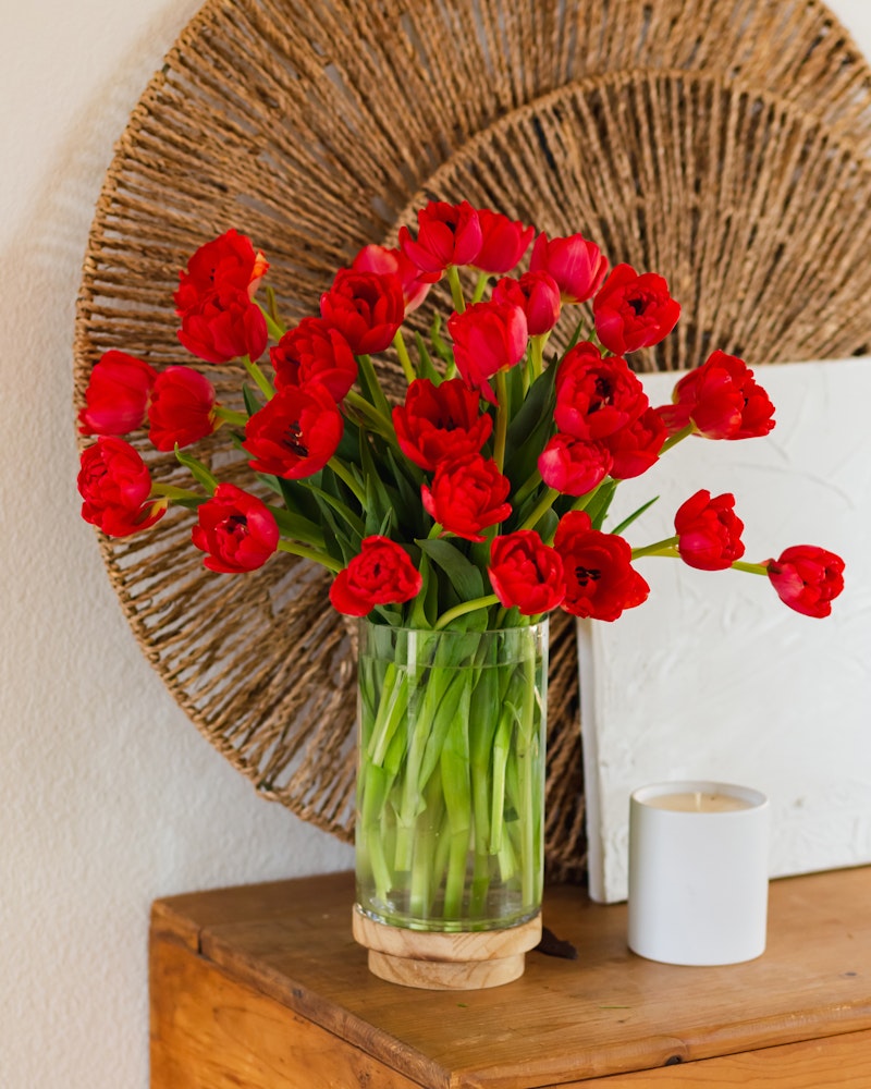 Vibrant red tulips arranged in a clear glass vase on a wooden side table, with decorative woven wall art in the background and a white candle to the side.