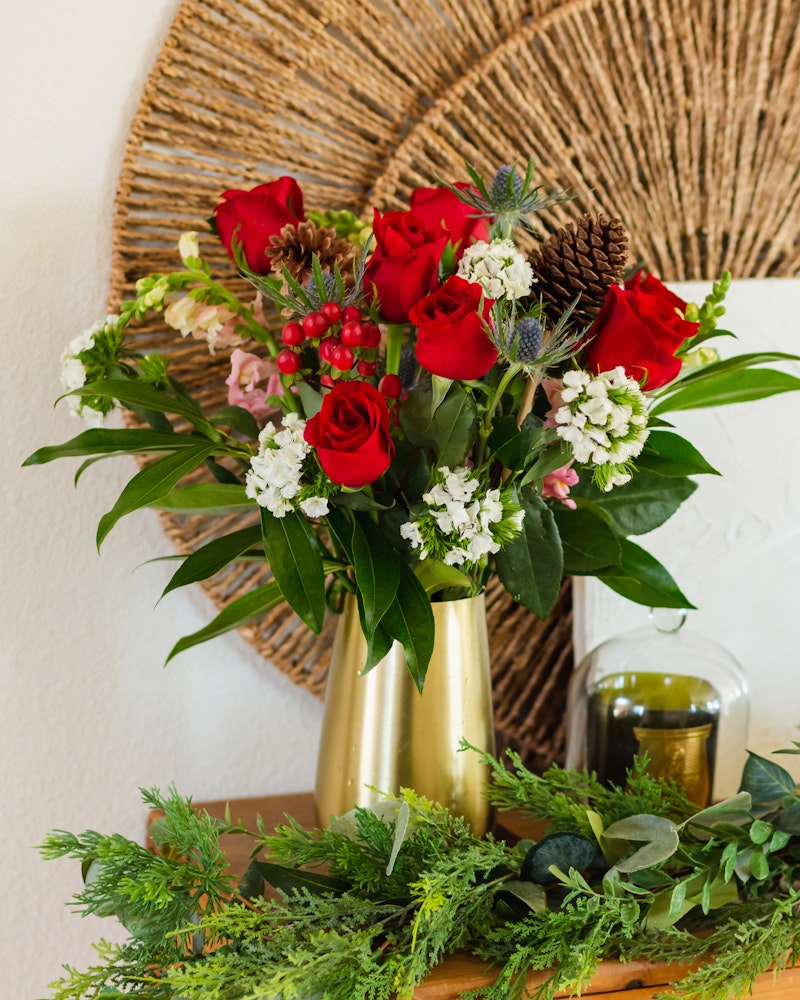 A vibrant bouquet of red roses and white flowers in a golden vase, with lush greenery on a wooden surface, set against a white wall and decorative wicker plate.