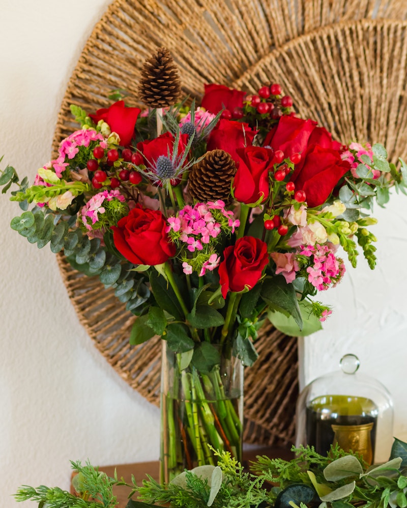 Lush bouquet of deep red roses highlighted with small pink blossoms and green foliage, paired with pine cones, set against a woven circular backdrop.