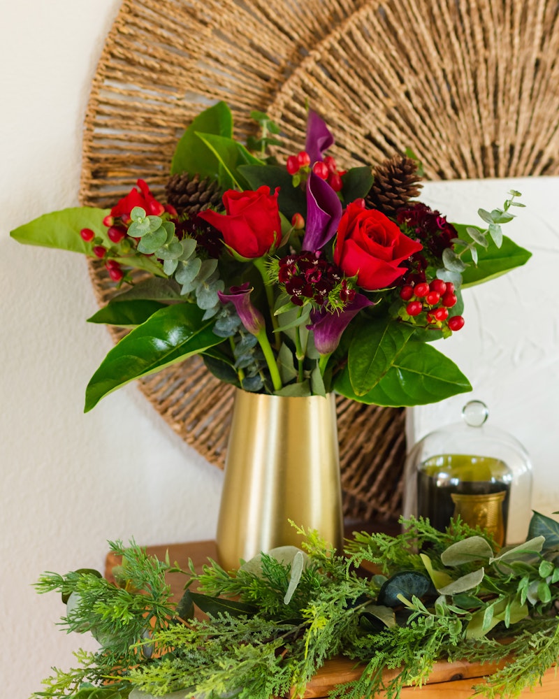 Vibrant bouquet of red roses and purple flowers in a gold vase, with green leaves and red berries, set against a textured wall with a circular woven decoration.
