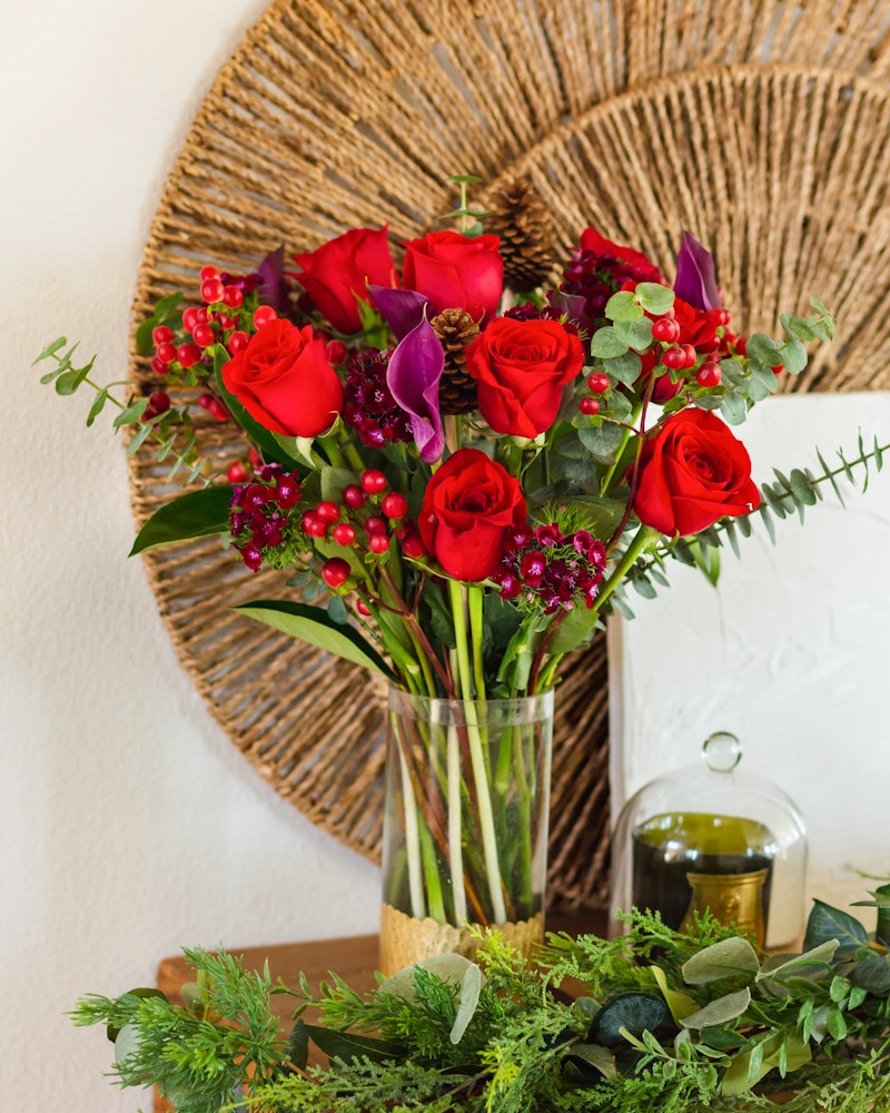 Vibrant bouquet of red roses and purple flowers arranged in a clear vase, with green leaves and red berries, set against a wicker plate backdrop on a white surface.