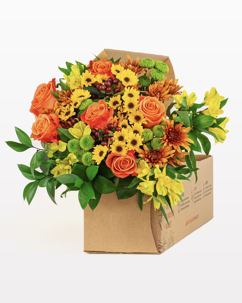 Vibrant bouquet of flowers with orange roses, yellow lilies, and greenery arranged in a cardboard box against a white background, ideal for gifts or decoration.