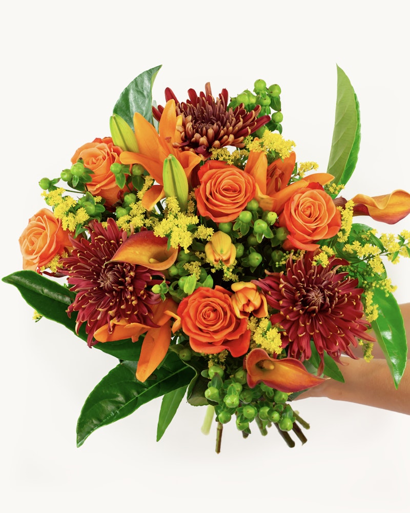 Person holding a vibrant bouquet with orange roses, lilies, and red chrysanthemums, accented by green foliage and yellow filler flowers against a white background.