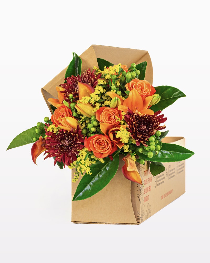 Vibrant bouquet of orange roses, red chrysanthemums, and yellow blooms with green foliage in a brown cardboard presentation box on a white background.