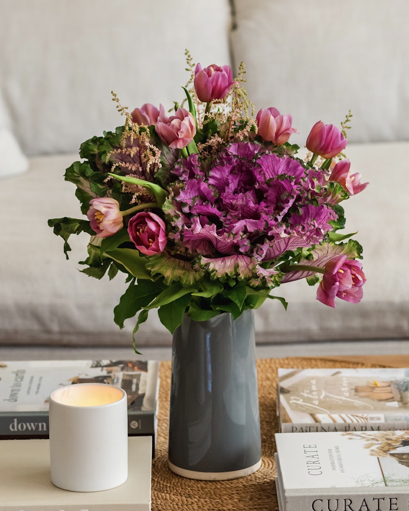 Vibrant bouquet of pink tulips and purple flowers arranged in a gray vase on a coffee table with magazines and a lit white candle, creating a cozy ambiance.