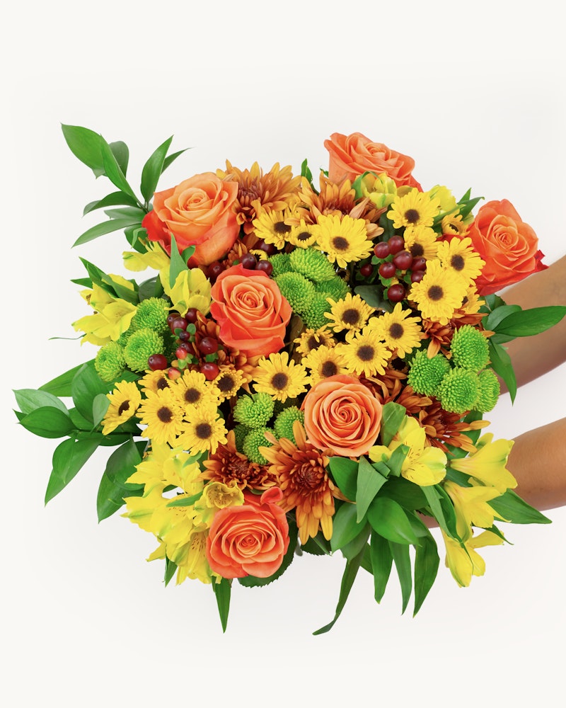 Two hands holding a vibrant bouquet of flowers including orange roses, yellow sunflowers, and green accents against a white background, conveying a fresh, springtime feel.