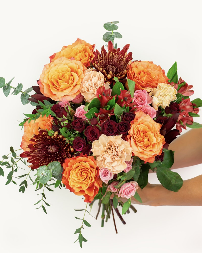 Vibrant bouquet of assorted flowers including orange roses, red chrysanthemums, and pink miniature roses held by hands against a white background.