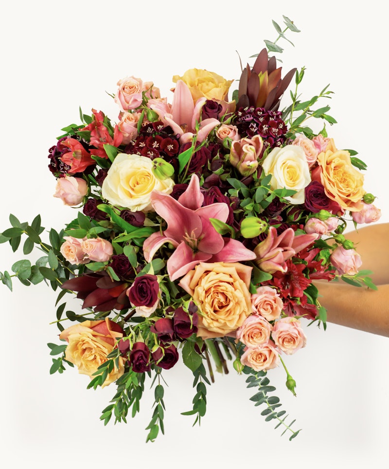 A vibrant bouquet of flowers including pink lilies, roses in shades of yellow and peach, deep red blooms, and lush greenery, held against a white background.