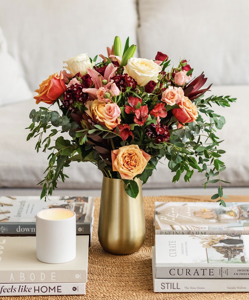 Lush bouquet of colorful roses and assorted flowers in a gold vase, on a table with books and a lit candle, creating a cozy home atmosphere.